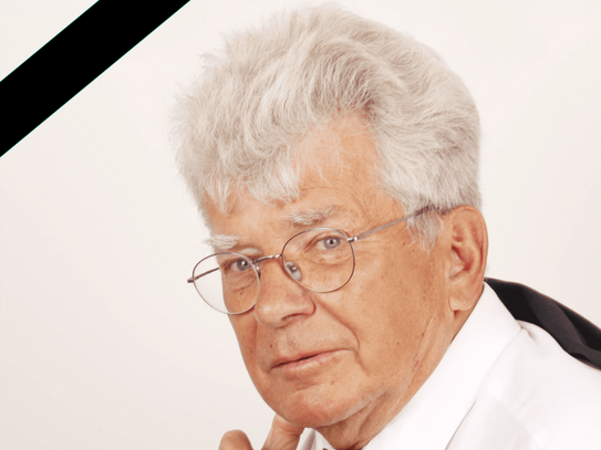 Porträt eines älteren Mannes mit grauen Haaren und Brille in weißem Hemd und roter Krawatte, nachdenklich blickend, mit schwarzem Trauerband in der oberen linken Bildecke.