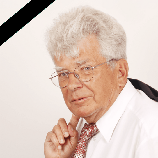 Porträt eines älteren Mannes mit grauen Haaren und Brille in weißem Hemd und roter Krawatte, nachdenklich blickend, mit schwarzem Trauerband in der oberen linken Bildecke.
