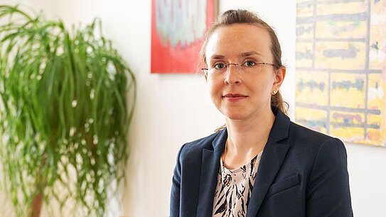 Frau im dunkelblauen Anzug sitzt auf einem hellbraunen Ledersofa in einem hellen Raum mit grüner Pflanze und bunten Gemälden im Hintergrund.