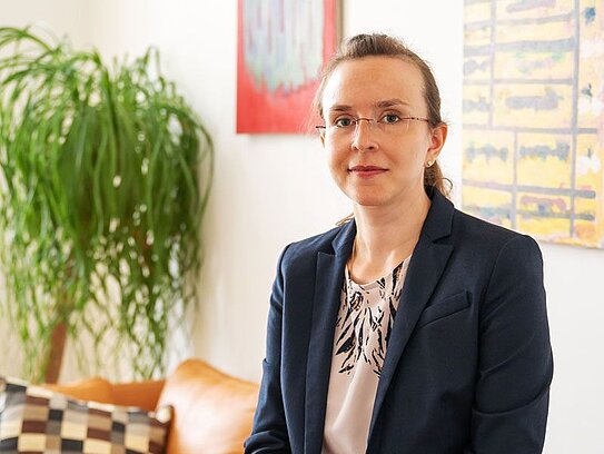 Frau im dunkelblauen Anzug sitzt auf einem hellbraunen Ledersofa in einem hellen Raum mit grüner Pflanze und bunten Gemälden im Hintergrund.