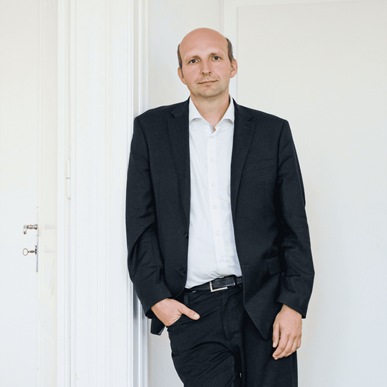 Ein Mann lehnt entspannt an einer weißen Wand in einem hellen Raum mit Holzboden. Er trägt einen schwarzen Anzug, ein weißes Hemd und schwarze Schuhe. Seine rechte Hand steckt in der Hosentasche, die linke hängt locker an seiner Seite. Er hat eine Glatze und blickt direkt in die Kamera. Im Hintergrund sind eine weiße Tür und eine Ecke des Raumes zu sehen.