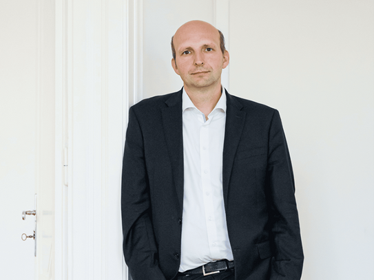 Ein Mann lehnt entspannt an einer weißen Wand in einem hellen Raum mit Holzboden. Er trägt einen schwarzen Anzug, ein weißes Hemd und schwarze Schuhe. Seine rechte Hand steckt in der Hosentasche, die linke hängt locker an seiner Seite. Er hat eine Glatze und blickt direkt in die Kamera. Im Hintergrund sind eine weiße Tür und eine Ecke des Raumes zu sehen.