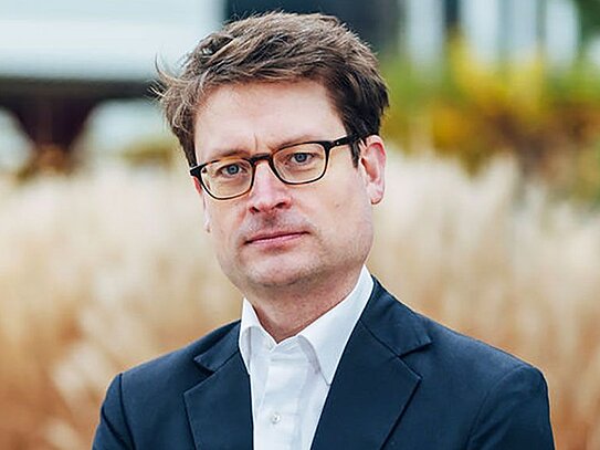 Porträt eines Mannes mit Brille und dunkelblauem Anzug vor unscharfem Pflanzen-Hintergrund.