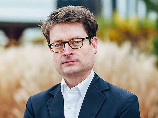 Porträt eines Mannes mit Brille und dunkelblauem Anzug vor unscharfem Pflanzen-Hintergrund.