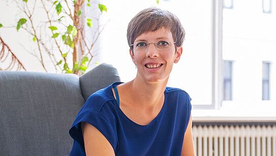 Frau mit kurzer brauner Frisur und Brille sitzt lächelnd in einem grauen Sessel, trägt blaue Bluse, schwarze Hosen und schwarze Sneaker, neben ihr ein Tisch mit Papieren und Smartphone, helle Innenraumbeleuchtung.