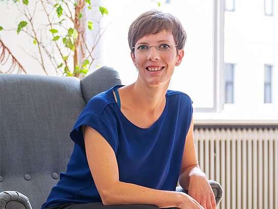 Frau mit kurzer brauner Frisur und Brille sitzt lächelnd in einem grauen Sessel, trägt blaue Bluse, schwarze Hosen und schwarze Sneaker, neben ihr ein Tisch mit Papieren und Smartphone, helle Innenraumbeleuchtung.