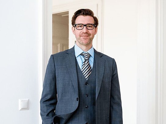 Mann im dunkelgrauen Anzug mit Weste, hellblauem Hemd und gestreifter Krawatte steht in einem hellen Raum mit Holzboden, lächelt und trägt eine Brille.