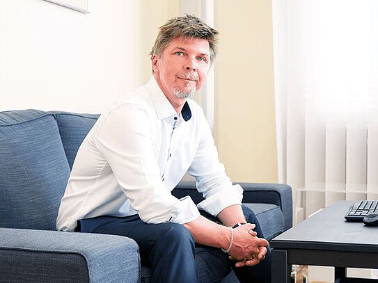 Ein Mann mittleren Alters mit kurzen, braunen Haaren und leichtem Bart sitzt auf einem dunkelblauen Sofa in einem hellen Raum. Er trägt ein weißes Hemd mit hochgekrempelten Ärmeln, eine dunkle Hose und schwarze Schuhe. Seine Hände sind vor ihm verschränkt, und er schaut leicht nach oben. Neben ihm steht ein schwarzer Couchtisch mit einer Tastatur und einer Computermaus darauf. Der Boden ist mit einem hellen, flauschigen Teppich bedeckt, und im Hintergrund sind helle Vorhänge vor einem Fenster zu sehen.