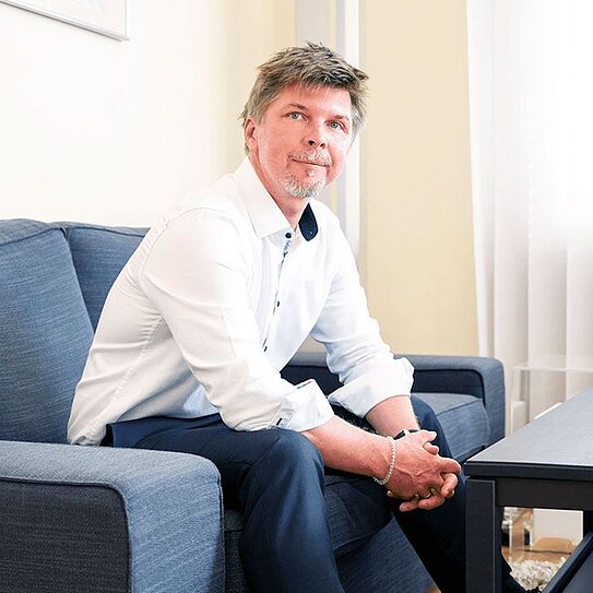 Ein Mann mittleren Alters mit kurzen, braunen Haaren und leichtem Bart sitzt auf einem dunkelblauen Sofa in einem hellen Raum. Er trägt ein weißes Hemd mit hochgekrempelten Ärmeln, eine dunkle Hose und schwarze Schuhe. Seine Hände sind vor ihm verschränkt, und er schaut leicht nach oben. Neben ihm steht ein schwarzer Couchtisch mit einer Tastatur und einer Computermaus darauf. Der Boden ist mit einem hellen, flauschigen Teppich bedeckt, und im Hintergrund sind helle Vorhänge vor einem Fenster zu sehen.